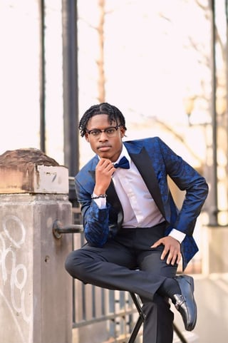 Young man in blue velvet blazer and bow tie sitting on concrete structure, posing thoughtfully outdoors
