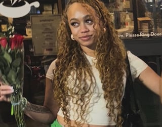 Woman with long curly blonde braids wearing a white crop top at an indoor venue with flowers in background