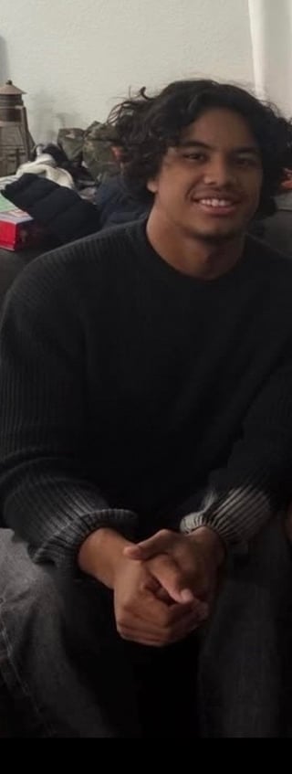 Young man with long dark hair wearing a black sweater, smiling at the camera while sitting indoors