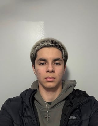Headshot of a young man wearing a black jacket and gray hoodie with a cross necklace, neutral expression against a white background