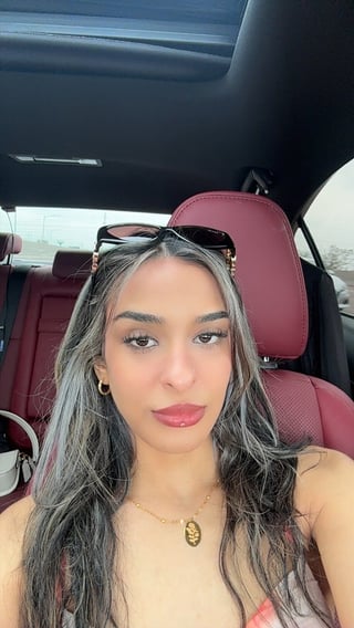 Woman with long dark hair and sunglasses on forehead taking a selfie inside a car with burgundy interior