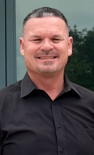 Man with short dark hair and facial hair wearing a black button-up shirt, smiling at the camera outdoors