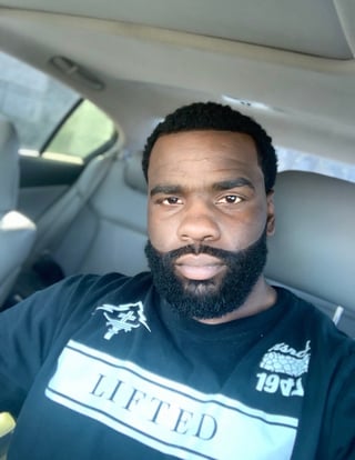 Man with dark hair and full beard wearing a black graphic t-shirt taking a selfie inside a car