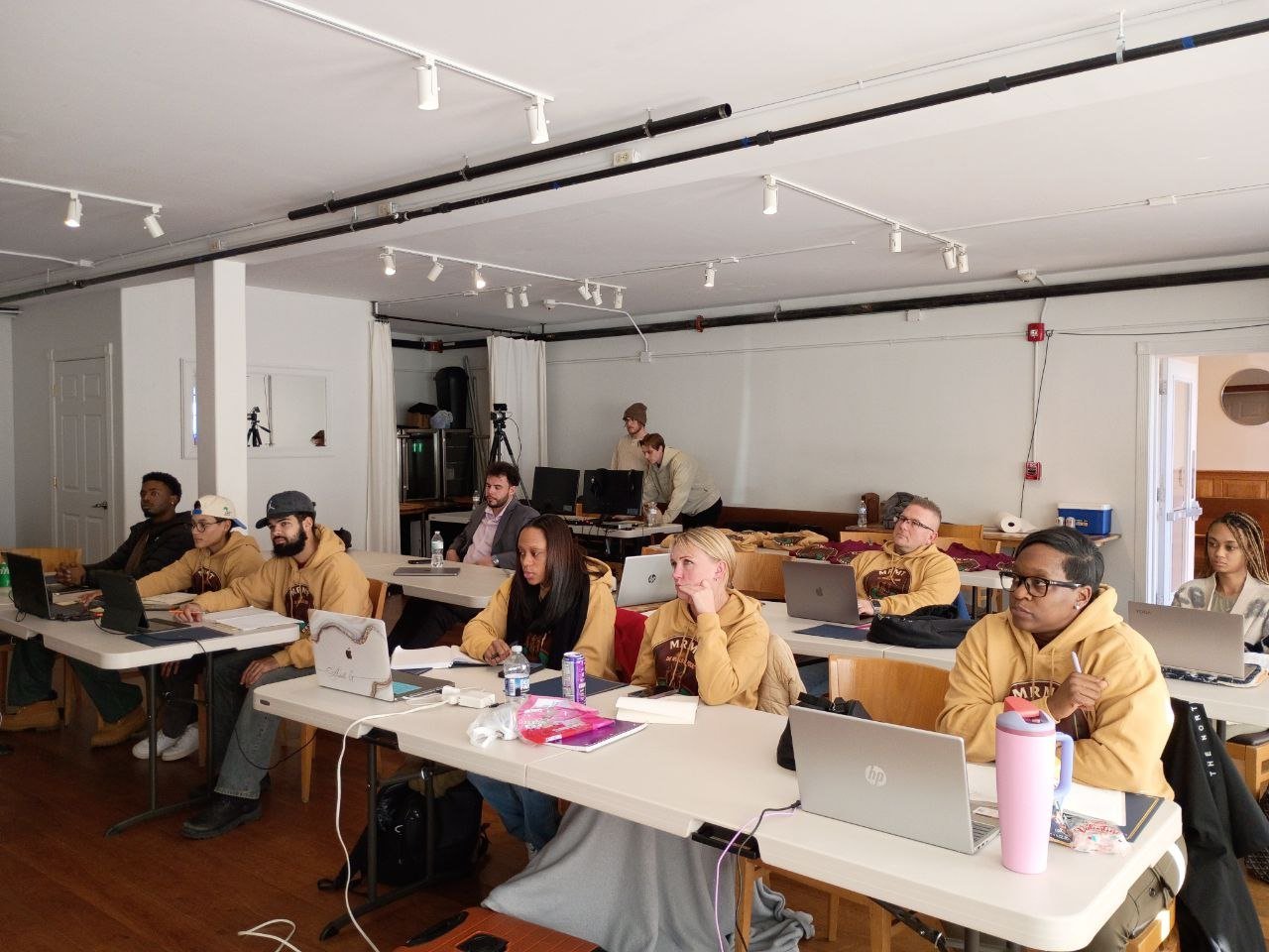 Group of people in yellow shirts seated at tables during a training or meeting event in a modern office space with track lighting