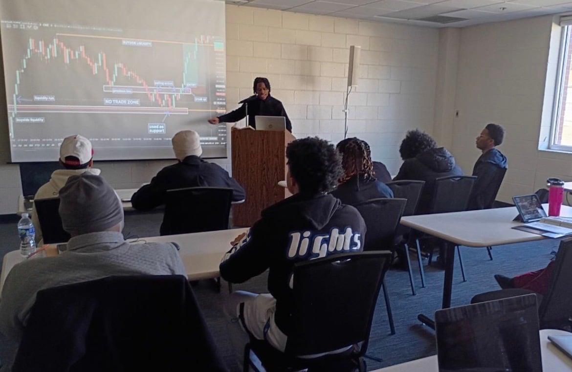 A presenter stands at a podium showing a financial chart on a projection screen to an audience seated in a classroom setting.